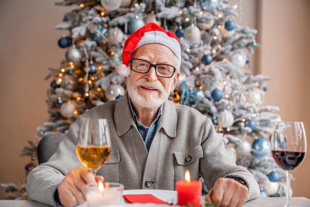 Oudere man in een kerstmanhoed die voor de kersttafel zit met een glas wijn