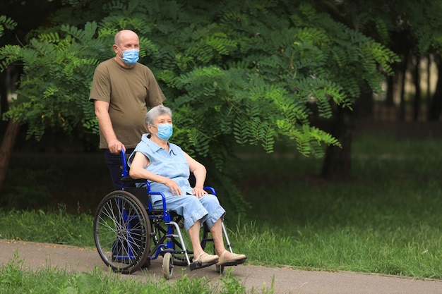 Oudere man lopen met gehandicapte bejaarde vrouw zittend in een rolstoel buiten het dragen van medische maskers