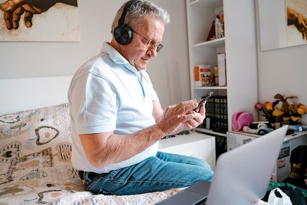 oudere man met draadloze koptelefoon die een mobiele telefoon vasthoudt met een laptop die thuis op het bed zit