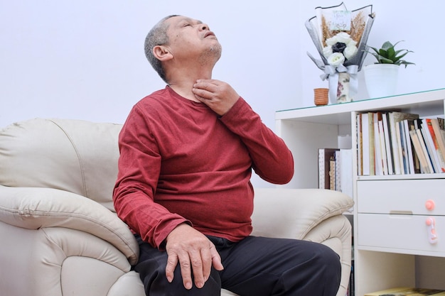 Oudere oudere man die op de bank zit en pijnlijke nek en keelpijn aanraakt voor griep, verkoudheid en infectie