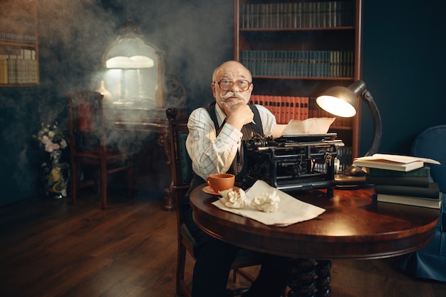 Oudere schrijver werkt aan vintage typemachine in zijn thuiskantoor. oude man met bril schrijft literatuurroman in kamer met rook