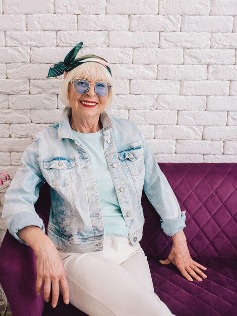 Foto oudere senior stijlvolle vrouw in blauwe zonnebril en spijkerjasje zitten op de bank in loft interieur