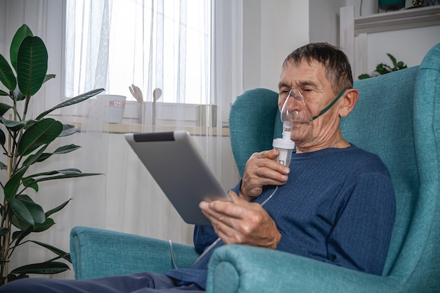 Oudere senior zit in een fauteuil met een zuurstofmasker en tablet