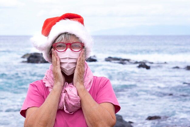 Oudere vrouw die een beschermend masker draagt vanwege de covid19 die op het strand staat met een kerstmuts