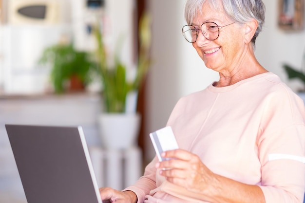 Oudere vrouw die op laptop surft en geniet van online winkelen Vrolijke en glimlachende mooie senior dame binnenshuis die in een goed humeur is met creditcard om te kopen