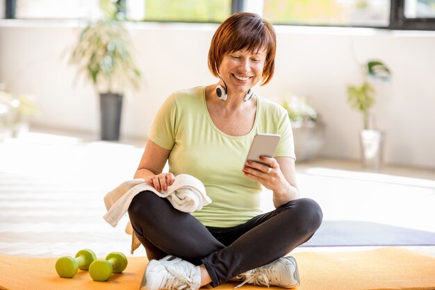 Oudere vrouw die smartphone gebruikt na sporttraining die binnenshuis of in de sportschool zit