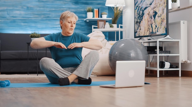 Oudere vrouw die thuis training op yogamat doet, na online trainingsles op laptop. Oudere persoon die fitnessvideo op de computer bekijkt om lichaamsbeweging op de vloer te doen.