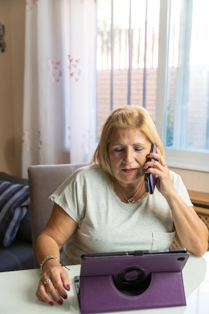 Oudere vrouw die thuis zit terwijl ze met iemand anders op haar mobiele telefoon praat en zichzelf vermaakt met haar tablet
