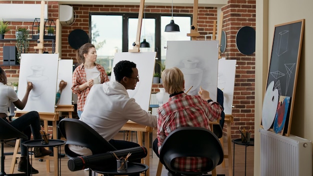 Oudere vrouw en jonge man leren schetsen op canvas te tekenen, praten in de kunstles. Team van studenten die samen een vaasmodel tekenen, waarbij ze kunstwerken gebruiken om de ontwikkeling van artistieke vaardigheden te oefenen.