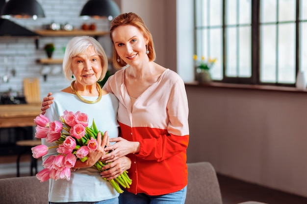 Oudere vrouw en schoondochter. Elegante verleidelijke gepensioneerde dame met roze tulpen in een stijlvolle blauwe top die wordt geknuffeld door een stralende, vrolijke, gracieuze roodharige volwassen schoondochter.