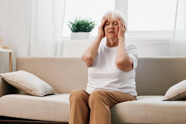 Oudere vrouw ernstige pijn in het hoofd zittend op de bank gezondheidsproblemen op oudere leeftijd slechte kwaliteit van leven Grootmoeder met grijs haar houdt haar hoofd vast migraine en hoge bloeddruk