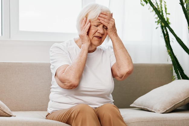 Oudere vrouw ernstige pijn in het hoofd zittend op de bank gezondheidsproblemen op oudere leeftijd slechte kwaliteit van leven Grootmoeder met grijs haar houdt haar hoofd vast migraine en hoge bloeddruk