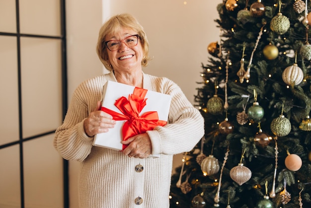 Oudere vrouw met een glimlach en een kerstcadeau bij een versierde boom