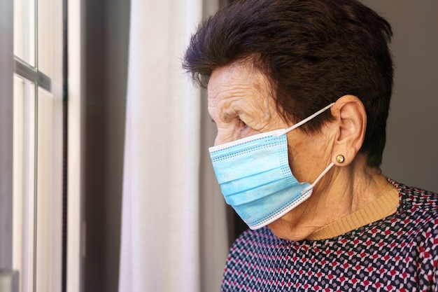 Oudere vrouw met een masker in haar huis kijkt uit het raam met angst voor de buitenkant, vanwege het coronavirus covid-19.