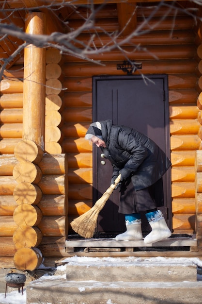 Oudere vrouw veegt sneeuw van veranda Blanke vrouw in zwarte kleding vilten laarzen en sjaal verwijdert sneeuw van veranda met bezem die viel na zware sneeuwstorm houten huisje
