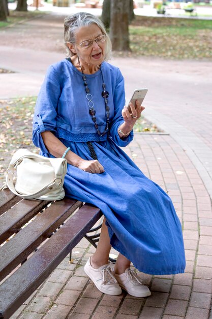 Oudere vrouw zit op de bank, communiceert via de telefoon in videochat en vertelt enthousiast iets