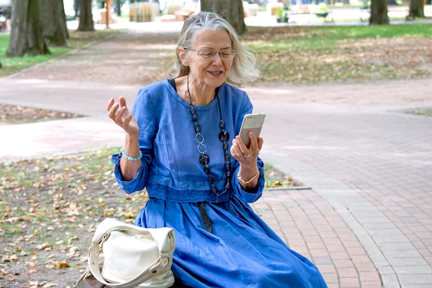 Oudere vrouw zit op een bankje in het park communiceert via de telefoon in videochat en vindt het leuk om iets te vertellen