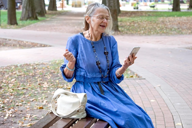 Oudere vrouw zit op een bankje in het park communiceert via de telefoon in videochat en vindt het leuk om iets te vertellen