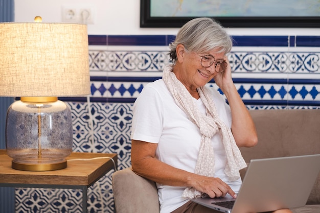 Oudere vrouw zittend op de bank tijdens het browsen op laptop Senior aantrekkelijke vrouw geniet van tech en social