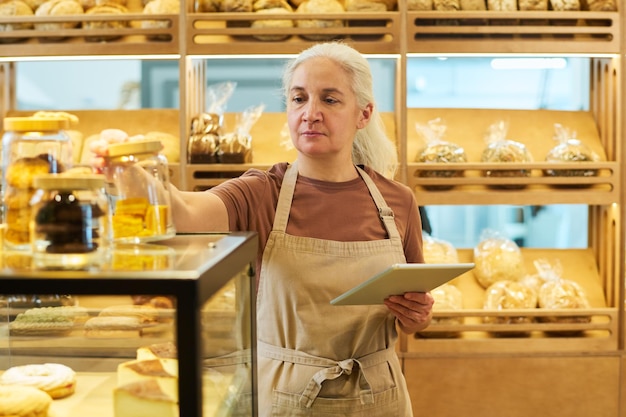 Oudere vrouwelijke eigenaar van een bakkerij met een tablet in de hand die bij de display staat