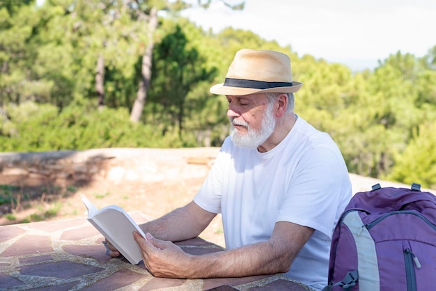 Oudere wandelaar zit aan een tafel buiten tussen de bomen en leest een boek