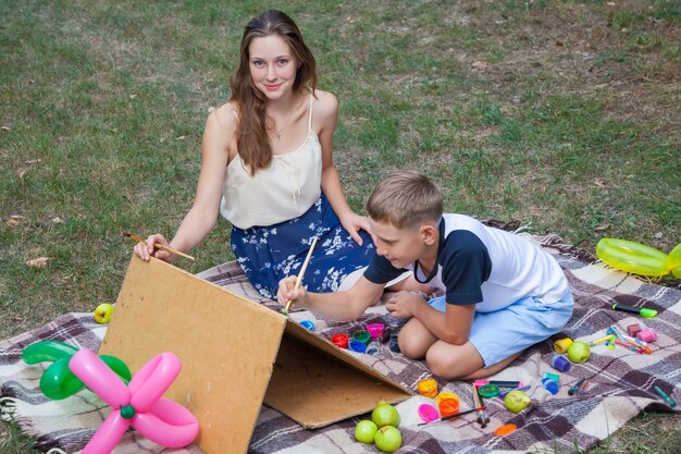 Oudere zus probeert haar broer te leren schilderen en poseren in het park in de zomer, kijkend naar de camera en glimlachen.