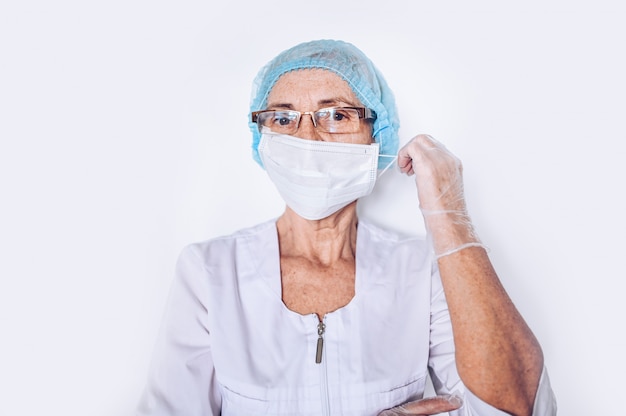 Ouderen triest moe volwassen vrouw arts of verpleegkundige in een witte medische jas, handschoenen, zet op gezichtsmasker het dragen van persoonlijke beschermingsmiddelen geïsoleerd. Gezondheidszorg en geneeskunde concept. Covid-19-pandemie