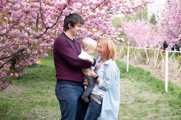 Ouders en zoontje van babyjongen die plezier hebben in het lentepark in de buurt van de roze sakura bloeiende boom Lenteconcept