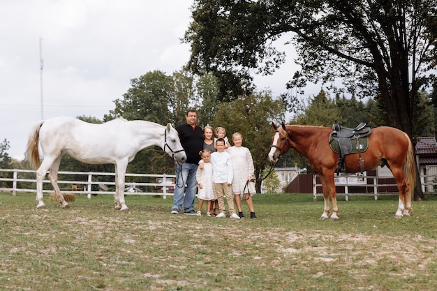 Ouders lopen met drie dochtertjes en zoon in de buurt van paarden op de boerderij op zomerdag. Vader en moeder brengen tijd door met kinderen op vakantie. Gelukkig gezin concept.