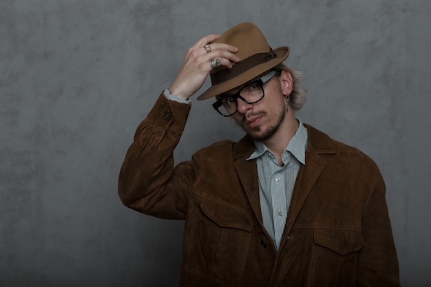 Ouderwetse jongeman hipster met baard trekt een stijlvolle hoed recht in een kamer bij een grijze muur.