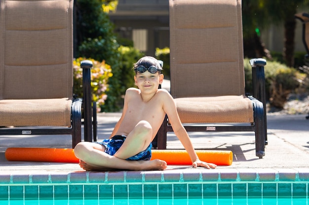屋外の夏のアクティビティ楽しい健康と休暇のコンセプト水泳メガネをかけた 8 歳の少年は、暑い夏の日にプールの近くに座っています。
