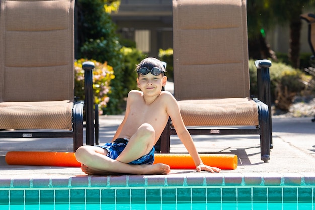 Oudoor summer activity Concept of fun health and vacation Boy eight years old in swim glasses sits near a pool in hot summer day