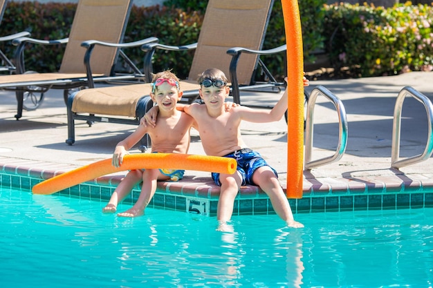 Photo oudoor summer activity concept of fun health and vacation brothers boys eight and five years old in swim glasses with noodles sits and laught near a pool in hot summer day