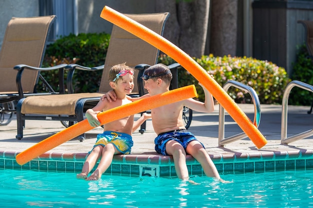 Photo oudoor summer activity concept of fun health and vacation brothers boys eight and five years old in swim glasses with noodles sits and laught near a pool in hot summer day