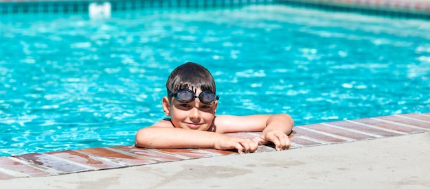 Foto attività estive all'aperto concetto di divertimento, salute e vacanza un ragazzo felice di otto anni con gli occhialini da nuoto si tiene sul lato della piscina in una calda giornata estiva