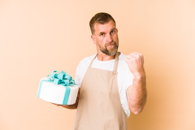 Oudste die een cake houdt die op beige muur wordt geïsoleerd die vuist, agressieve gezichtsuitdrukking toont.