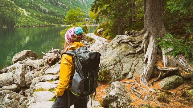 Oung blond hair woman with big camping rucksack wearing yellow raincoat jacket purple and blue cap looking on mountains and lake Copy space rear view background