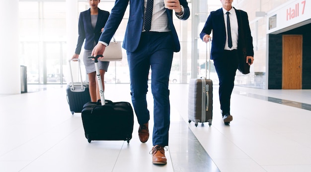 La nostra attività ci richiede di viaggiare immagine ritagliata di tre uomini d'affari irriconoscibili che camminano e tirano le valigie mentre sono in ufficio durante il giorno