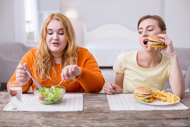 Our lunch together. Serious slim woman eating fast food and talking with her fat friend eating a salad