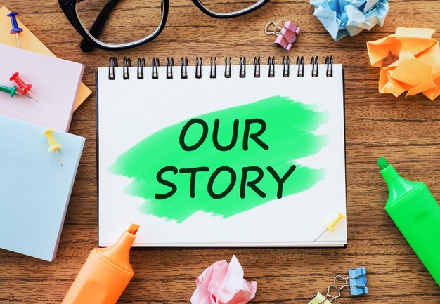 OUR STORY text on notepad on wooden table with open diary, glasses, markers, crumpled paper and paper clips