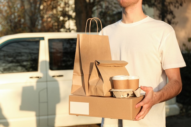 Сourier holds blank box, coffee cups and paper packages against car