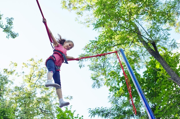 Outdoor active recreation and amusement park, child girl having\
fun jumping on trampoline with elastic ropes