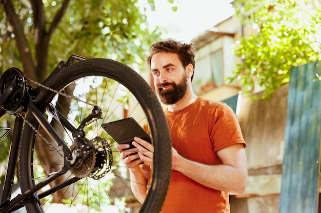 屋外でアクティブな活気に満ちた白人男性が、自転車のメンテナンスのためにインターネットを探索しています。電話タブレットを利用して、夏のレジャーサイクリングのために自転車の車輪を修理する、熱心な健康な男性サイクリスト。