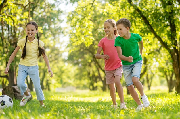 Outdoor activities. Two smiling cute junior girls and joyful boy chasing ball together on grass in park