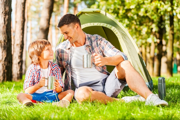 Outdoor adventure. Cheerful father and son sitting near the tent and drinking hot tea from metal cups while camping in the forest together