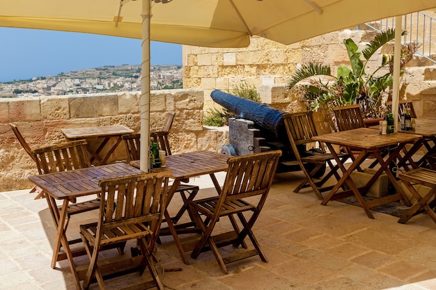 An outdoor cafe terrace with tables and chairs with a view of a coastal city on a sunny day.