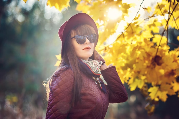 Outdoor primo piano ritratto modello di grandi dimensioni bella ragazza bruna con cappello e occhiali da sole guardando la fotocamera nella foresta autunnale come sfondo