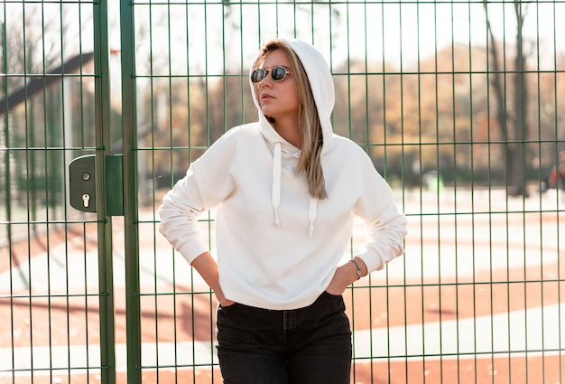 Ritratto alto vicino all'aperto di giovane bella donna con capelli lunghi in occhiali da sole, vestito con un maglione con cappuccio bianco, vicino al campo sportivo. cultura giovanile passatempo estivo