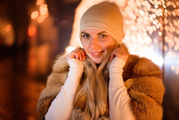 Внешний конец вверх по портрету молодой счастливой усмехаясь девушки представляя на улице. Снегопад, праздничная гирлянда.