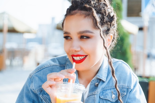 Outdoor closeup portrait of a cute stylish fashionable girl having fun drinking orange juice in an outdoor cafe a woman with dreadlocks and red lipstick